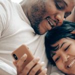 Communication - Cheerful young diverse couple lying together in bed and smiling while watching video on mobile phone in morning