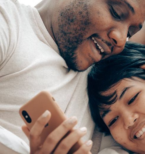 Communication - Cheerful young diverse couple lying together in bed and smiling while watching video on mobile phone in morning