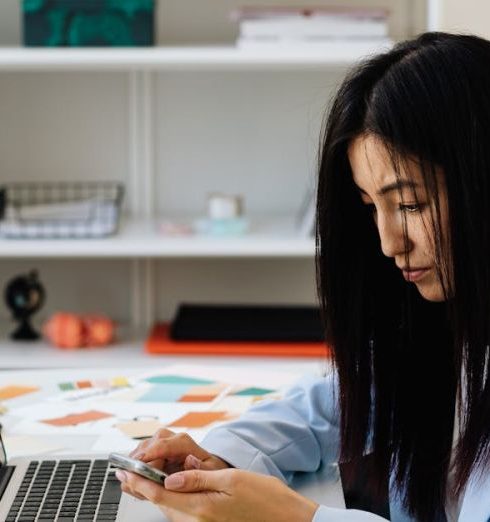 Emails - Professional woman using laptop and smartphone in modern office setting.