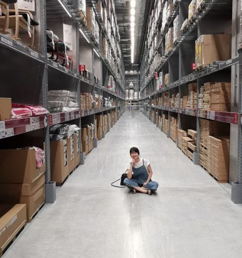 Inventory - An Asian woman sits in a large warehouse aisle filled with inventory racks and shelves.