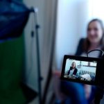 Video Marketing - Woman being recorded in a professional studio setup, using video camera and lighting equipment.