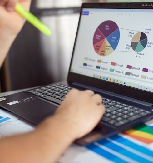 Sales Metrics - Person analyzing financial charts and graphs on a laptop with colorful documents, showcasing market analysis.