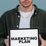 Lead Generation - Adult wearing casual clothing holding a tablet that reads 'Marketing Plan'.