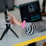 Personal Brand - Woman adjusting smartphone on tripod with laptop setup for video editing.