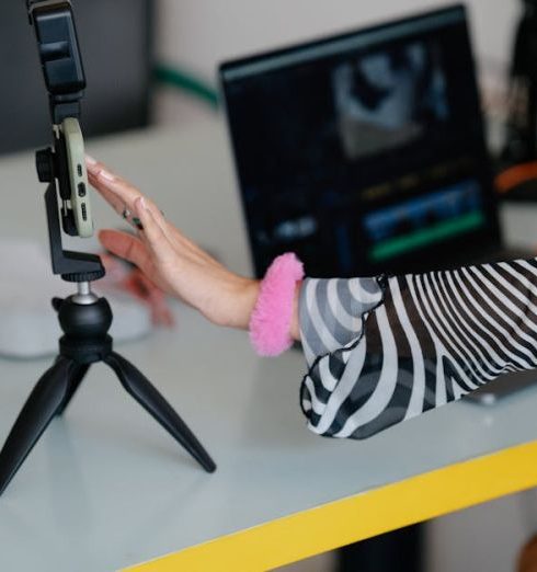 Personal Brand - Woman adjusting smartphone on tripod with laptop setup for video editing.