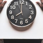 Time Management - High angle view of a man with an apple watch observing a clock on a white table.