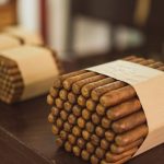 Product Bundles - Stacks of cigar bundles wrapped in paper, placed on a wooden surface inside a manufacturing facility.