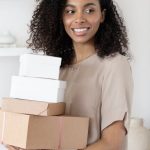 Account-Based Selling - Young African American woman smiling while holding cardboard boxes indoors.