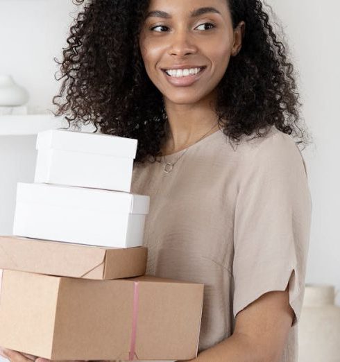 Account-Based Selling - Young African American woman smiling while holding cardboard boxes indoors.