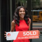 Sales Confidence - Smiling woman in red dress holding sold sign outside an office building.