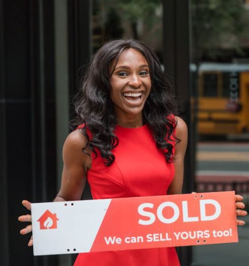 Sales Confidence - Smiling woman in red dress holding sold sign outside an office building.