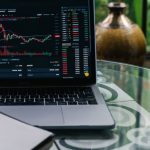 Dividend Stocks - Laptop displaying cryptocurrency stocks and graphs on a glass table with a notepad.