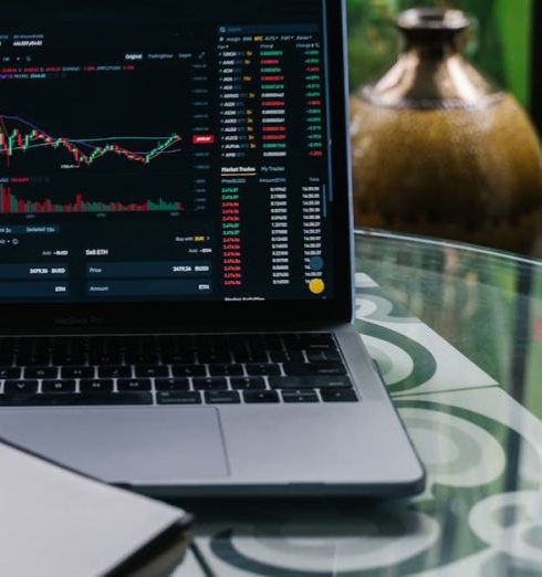 Dividend Stocks - Laptop displaying cryptocurrency stocks and graphs on a glass table with a notepad.