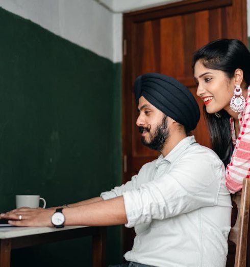 Home Office - Side view of positive young Sikh man in shirt and turban working on laptop at home while wife leaning on chair behind