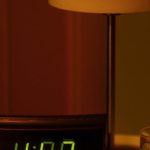 Paid Time Off - A warm and moody bedside table with a digital clock showing 4:08 and a lamp in dim lighting.