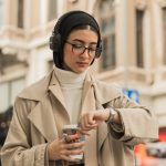 Timekeeping - Young woman with a headscarf checks her watch while holding coffee on an urban street.