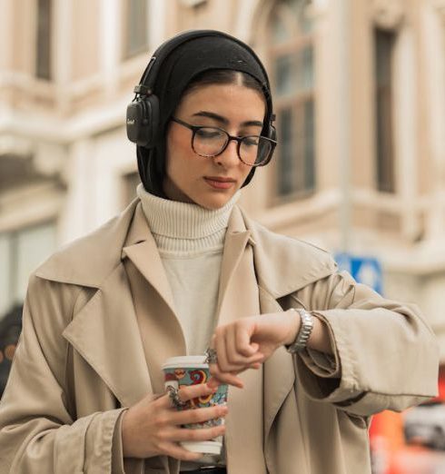 Timekeeping - Young woman with a headscarf checks her watch while holding coffee on an urban street.