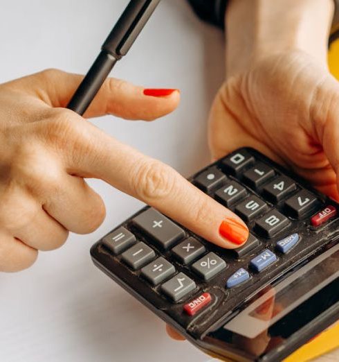 Metrics - Close-up of a woman using a calculator at her wooden workspace with documents and notepads.