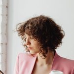 Effectiveness - Elegant woman in a pink blazer texting on her smartphone while holding a coffee cup indoors.