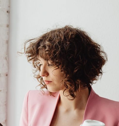 Effectiveness - Elegant woman in a pink blazer texting on her smartphone while holding a coffee cup indoors.