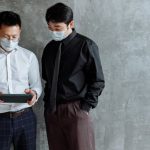 Contingency Plan - Two men in business attire and face masks using a tablet indoors.