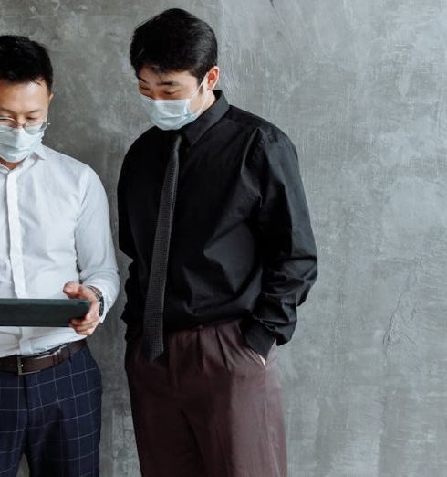 Contingency Plan - Two men in business attire and face masks using a tablet indoors.