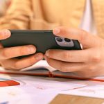 Strategy Updates - Woman Sitting at a Table and Using Smart Phone with an Open Notebook