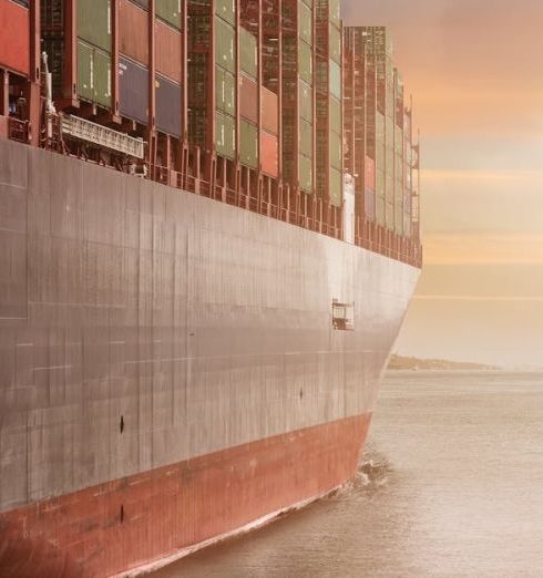 Supply Chain - A colossal cargo ship loaded with containers navigates through calm waters against a vibrant sunset sky.