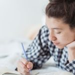 SWOT Analysis - Young woman in checkered pajamas writing in a notebook while lying on a bed.