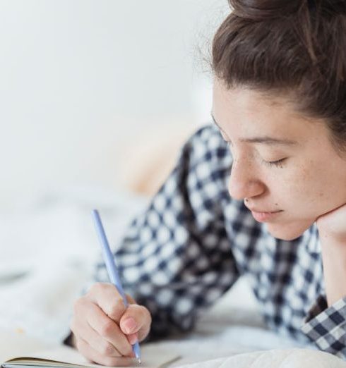 SWOT Analysis - Young woman in checkered pajamas writing in a notebook while lying on a bed.