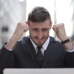 Purpose - A joyful businessman in a suit triumphantly celebrates success on his laptop outdoors.