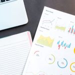 Data Visualization - Flatlay of business report with colorful charts, a notebook, and a laptop for data analysis on a desk.