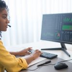 Customer Analytics - African American woman working in office analyzing data on dual computer monitors.