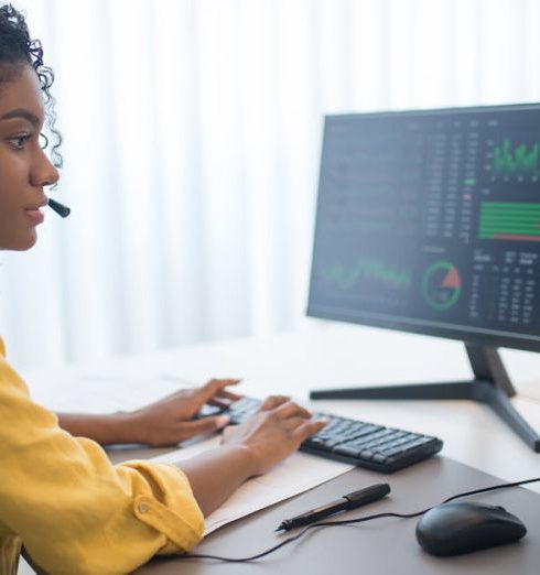 Customer Analytics - African American woman working in office analyzing data on dual computer monitors.