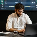 Marketing Analytics - A focused man analyzing stock market trends on a laptop in an office setting with charts displayed.