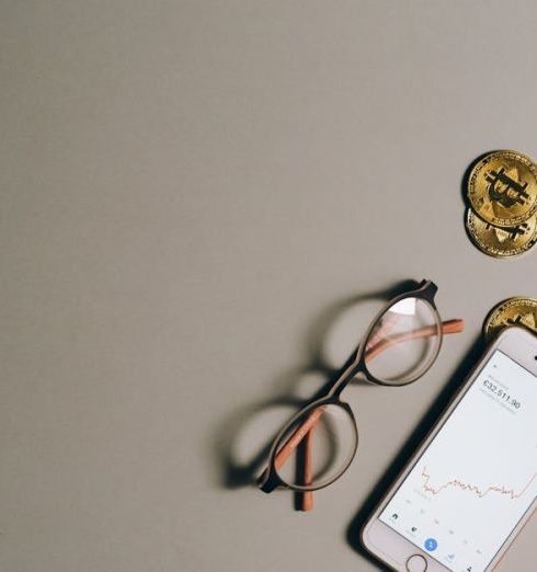 Financial Analytics - Flat lay of Bitcoin coins, smartphone with graph, and glasses on gray surface.