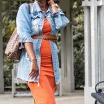 Benchmarking - Stylish woman in orange dress and denim jacket posing in a park gazebo during fall.