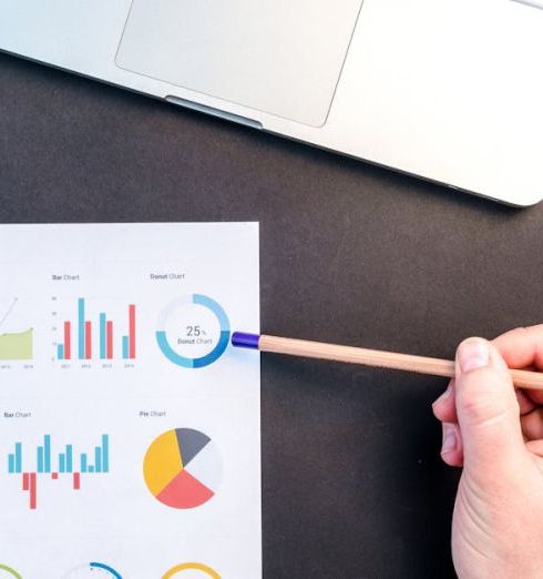 Market Research - Hand holding pencil reviewing colorful data charts on desk with laptop.