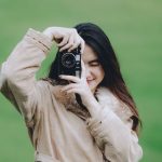 Local Talent - A young woman in a coat takes a photo with a camera outdoors on a green field.