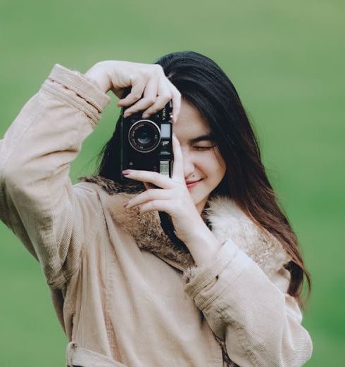 Local Talent - A young woman in a coat takes a photo with a camera outdoors on a green field.