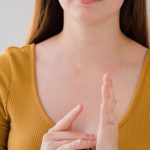 Language Barriers - Portrait of a young woman gesturing in sign language, wearing a yellow top and looking at the camera.