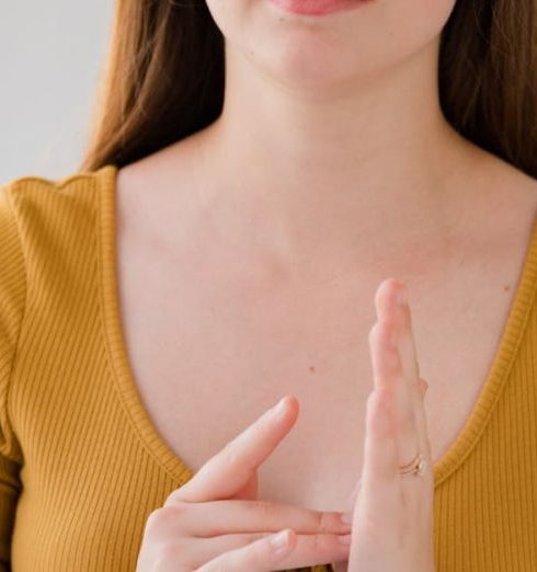 Language Barriers - Portrait of a young woman gesturing in sign language, wearing a yellow top and looking at the camera.