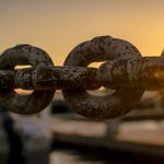 Supply Chain - Close-up of a rusty chain with sunrise and water reflections in the background.