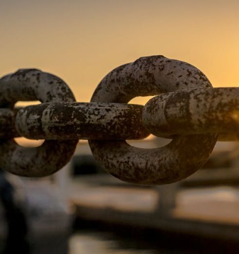 Supply Chain - Close-up of a rusty chain with sunrise and water reflections in the background.