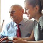 Customer Support - Young woman in casual clothes helping senior man in formal shirt with paying credit card in Internet using laptop while sitting at table