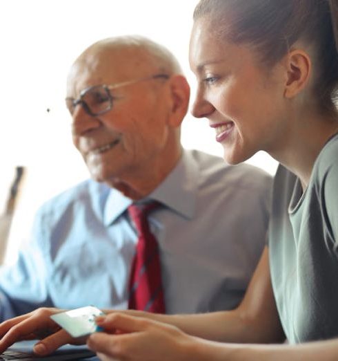 Customer Support - Young woman in casual clothes helping senior man in formal shirt with paying credit card in Internet using laptop while sitting at table