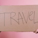 Cross-Border - Two hands holding a cardboard sign with 'TRAVEL' on a pink backdrop.