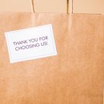 Brand Loyalty - A hand presents a brown paper shopping bag with a thank you note on a warm background.