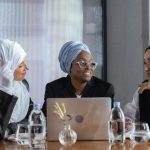 Cross-Cultural Collaboration - Three diverse women in hijabs discuss ideas around a table with a laptop, promoting inclusivity.