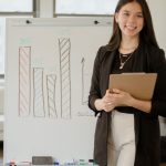 Success Metrics - Confident businesswoman presenting charts and data analysis on a whiteboard in a modern office setting.